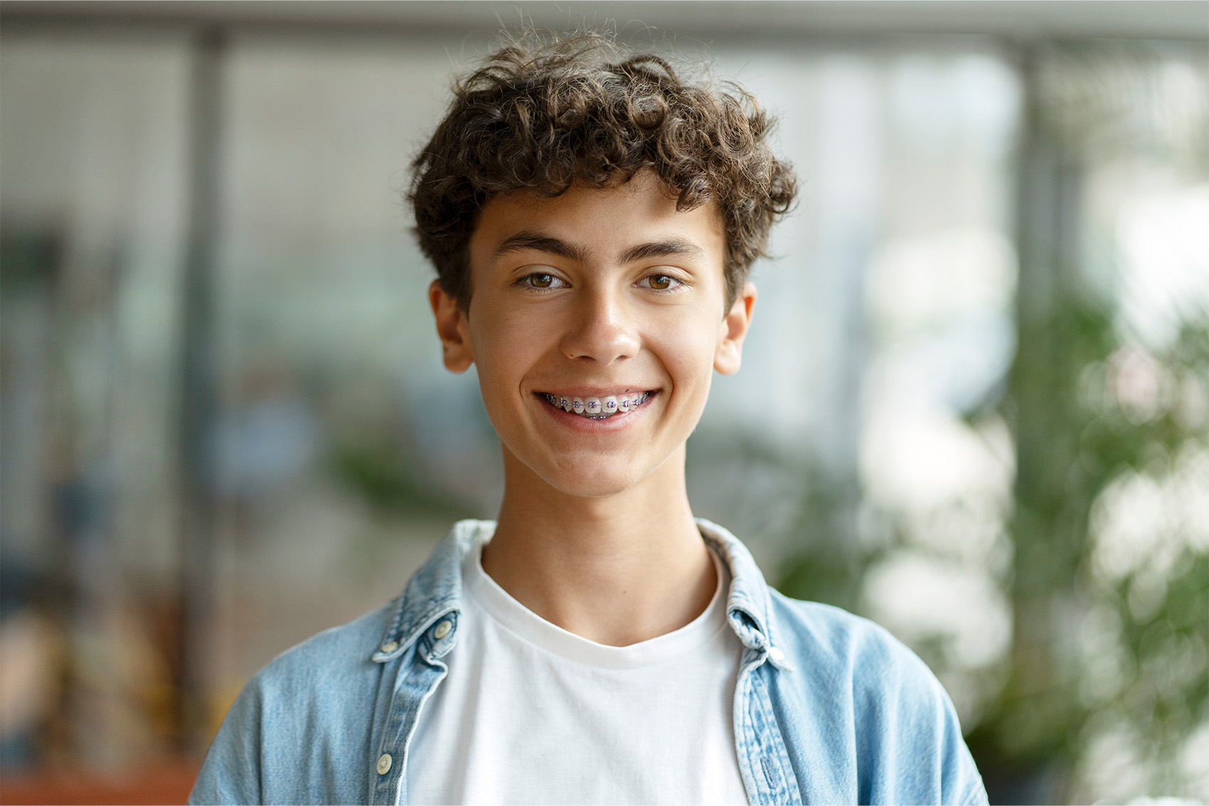 A young man with curly hair smiles, showcasing his braces in a dental office setting.